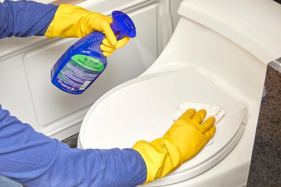 A woman wearing rubber gloves using spray cleaner to clean the outside of a toilet.