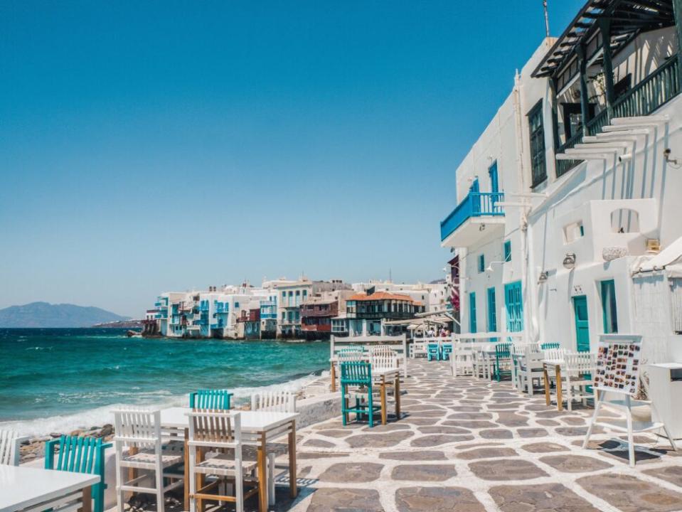 Pictured: Sunny Mykonos coastline with colorful white and blue buildings, stone walkway, and restaurant chairs by the water