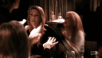 Two women, Kim Richards and Lisa Rinna, are in a heated argument at a restaurant table. Kim is seated, gesturing with her hand, and Lisa appears upset