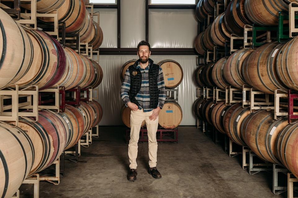 Man standing in between rows of wooden wine barrels