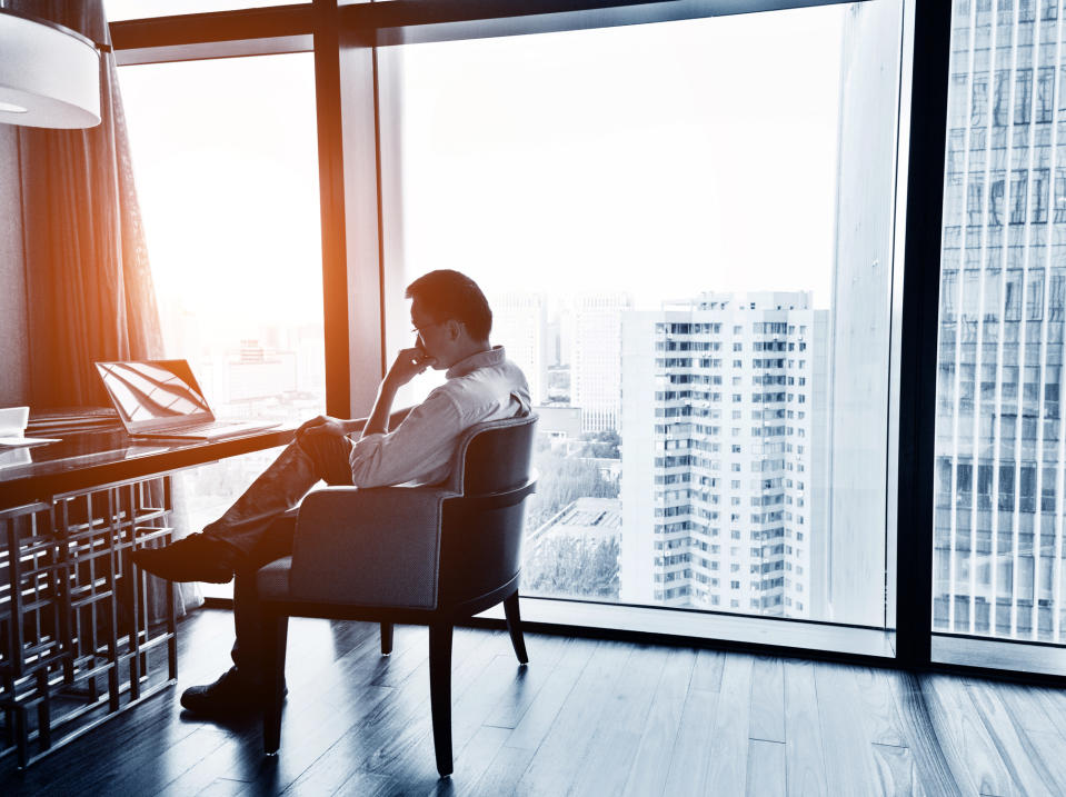 A man in an office sits at a desk with a laptop, looking thoughtful while gazing out a large window at a cityscape