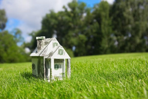 A house made of folded U.S. dollar bills sits on a grassy lawn.