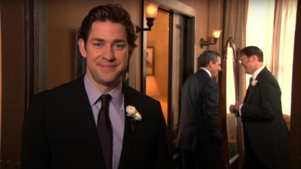 John Krasinski smiles as Steve Carell and Rainn Wilson talk in the background in The Office.