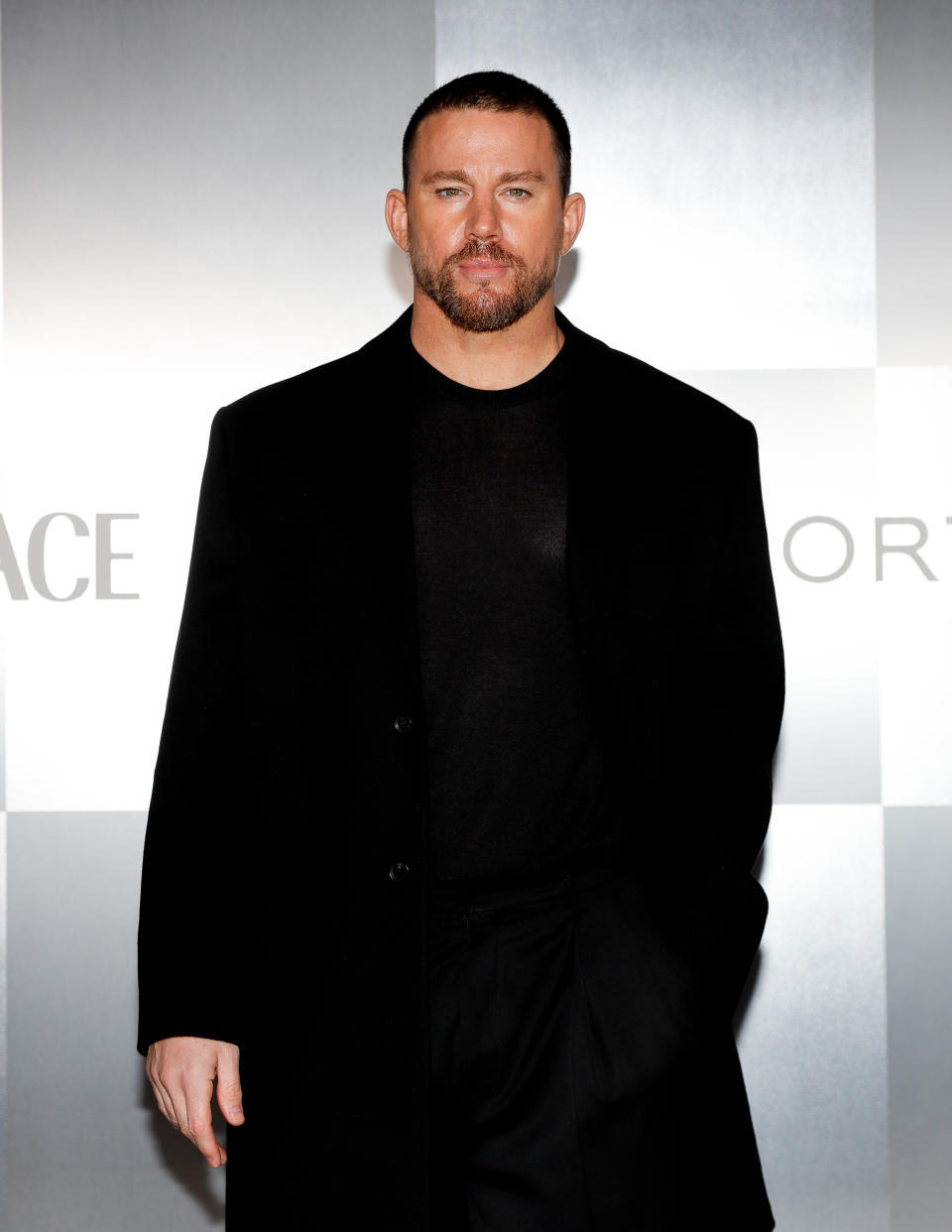 Channing Tatum is on a red carpet wearing a black coat over a black shirt and pants, against a backdrop with text "Versace."