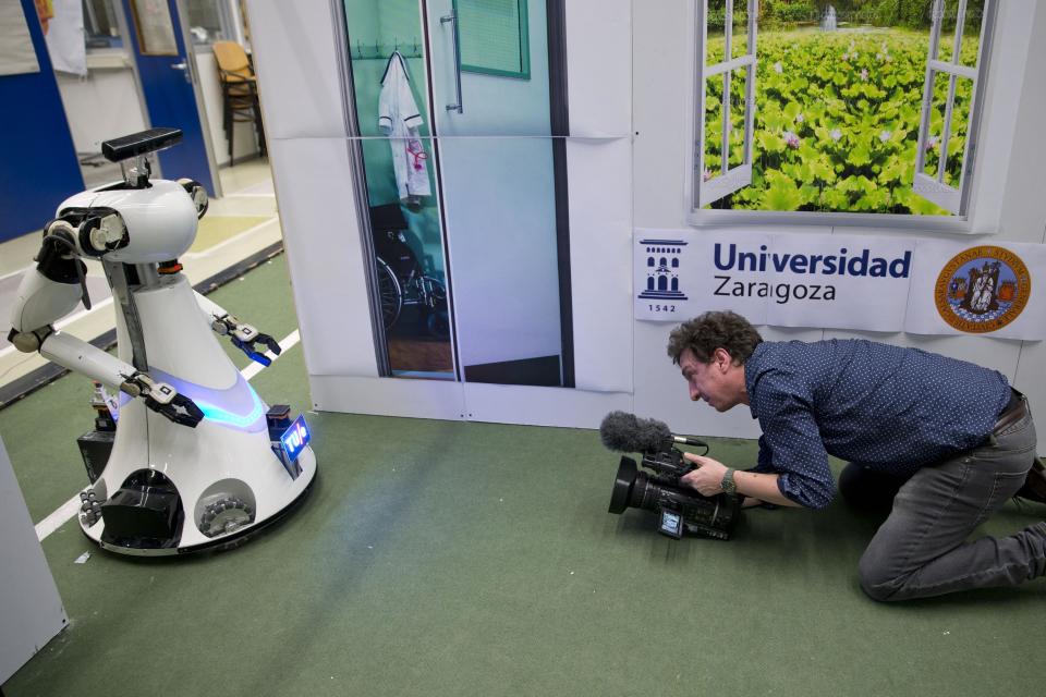 A cameraman films Amigo, a white robot the size of a person, who uses information gathered by other robots to move towards a table to pick up a carton of milk and deliver it to an imaginary patient in a mock hospital room at the Technical University of Eindhoven, Netherlands, Wednesday Jan. 15, 2014. A group of five of Europe's top technical universities, together with technology conglomerate Royal Philips NV, are launching an open-source system dubbed "RoboEarth" Thursday. The heart of the mission is to accelerate the development of robots and robotic services via efficient communication with a network, or "cloud". (AP Photo/Peter Dejong)