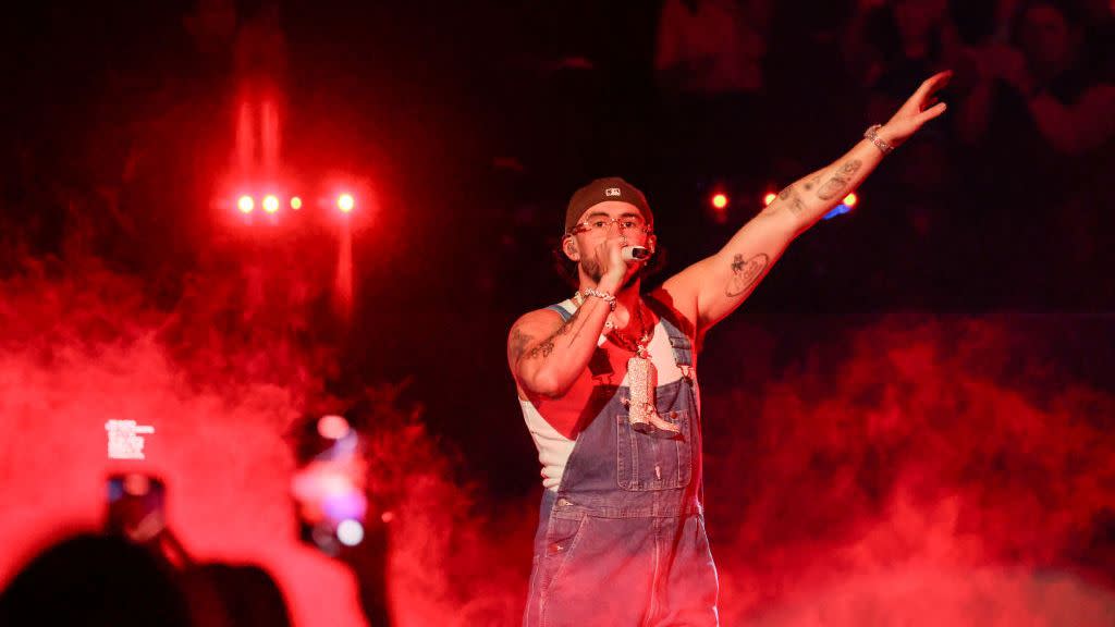 bad bunny holds a microphone to his mouth and stands on a stage with his other arm extended into the hair, he wears overalls and a white tank top with a backward ball cap and glasses