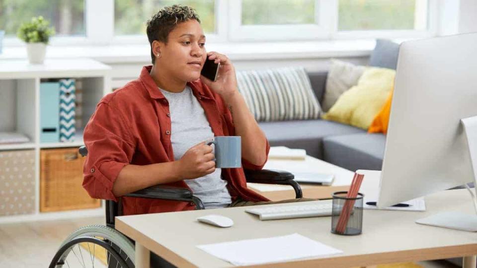 woman sitting in wheelchair at the table and looking at computer monitor while talking on mobile phone and drinking coffee at home