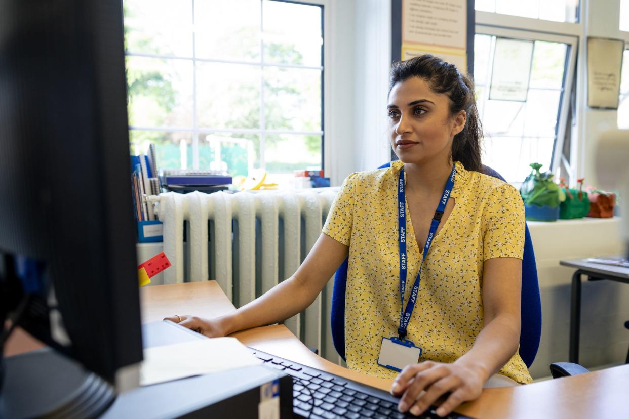 A close up front view of a young teacher sitting at her desk in her classroom. She is checking her lesson plans on her desktop PC. She is working in a school in Hexham in the North East of England.