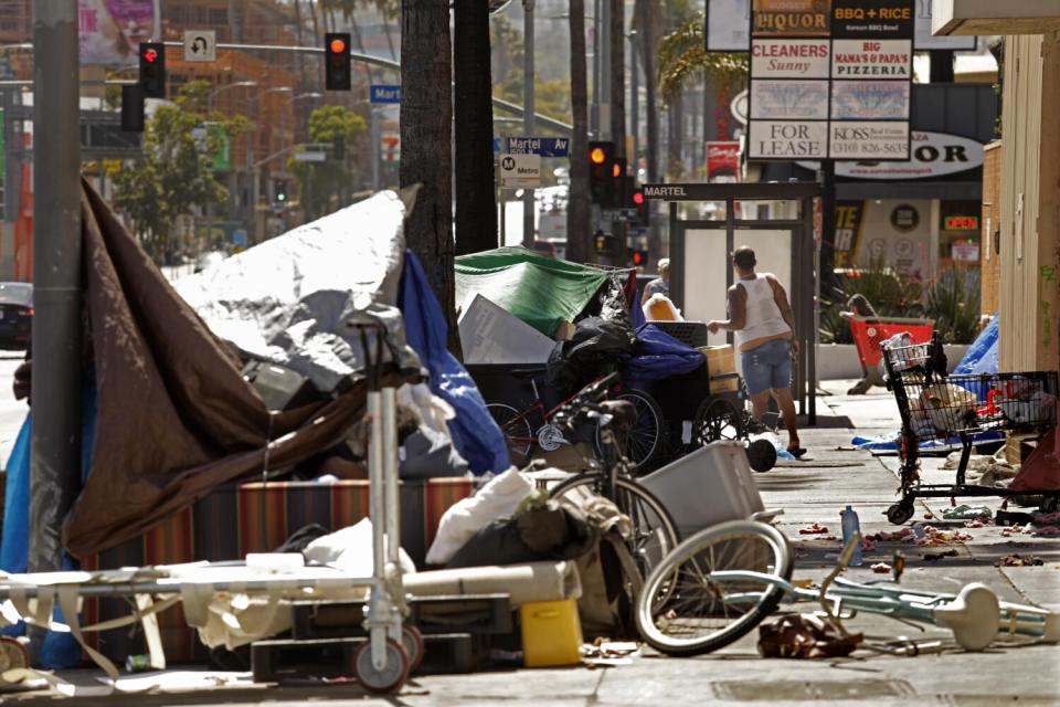 A homeless woman stands in the middle of an encampment on the sidewalk along a busy street