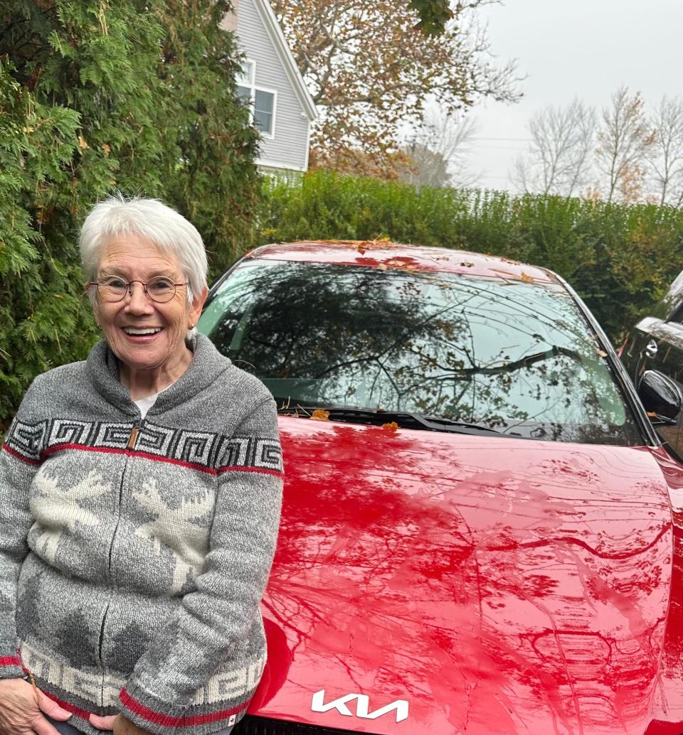 A woman in a sweater stands in front of her red car.