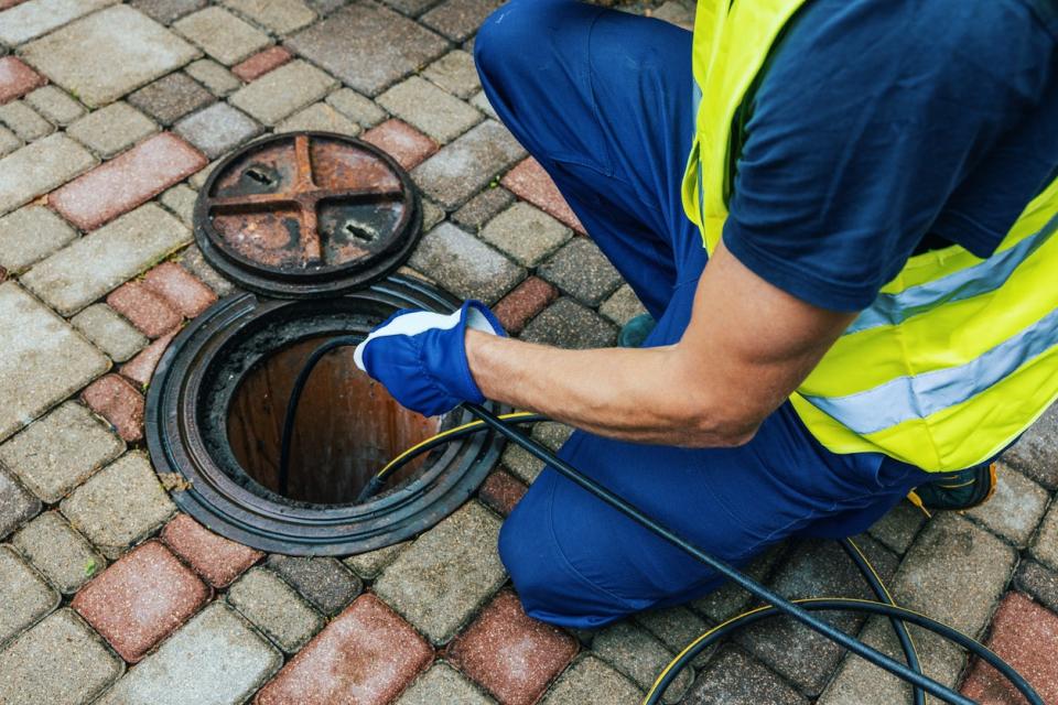 A close up of a worker in a blue work-suit and yellow vest assess underground plumbing. 