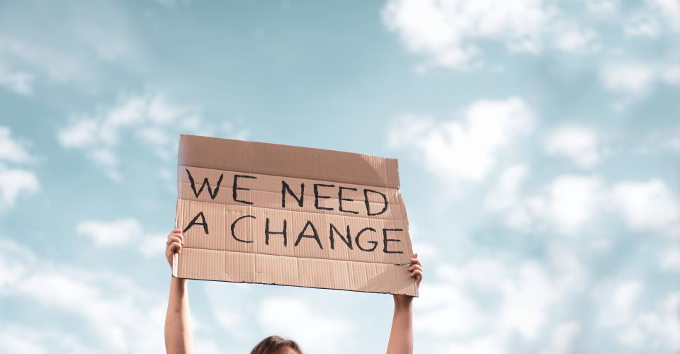 Woman holding a banner of 'We Need A Change'. Young woman with poster in front of people protesting about climate changing on the street. Meeting about problem in ecology, environment, global warming, industrial influence, climate emergency.