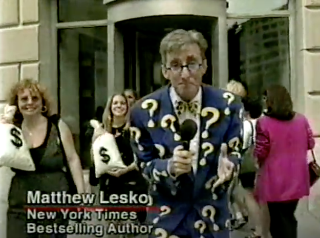 Matthew Lesko in a question mark suit holds a book and microphone outside a building