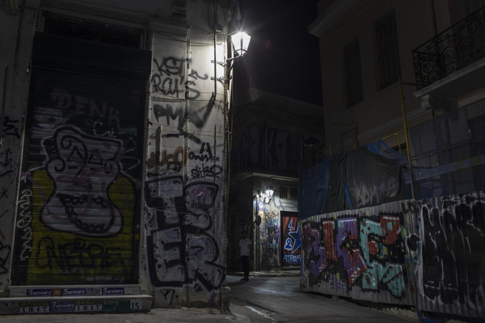 In this Monday, July 22, 2019 photo, a man stands in a street next to a shop and houses covered with graffiti in Athens' Plaka district. Some parts of central Athens are so afflicted with graffiti _ largely undecipherable squiggles in bold, broad strokes _ that few facades remain untouched and property owners give up on repainting. (AP Photo/Petros Giannakouris)