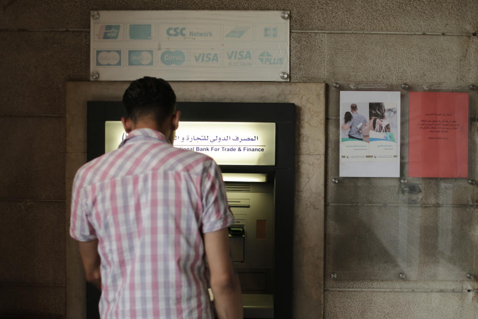 FILE - This July 24, 2019 file photo, a man stands in front of an ATM machine outside a branch of the International Bank for Trade and Finance, in Damascus, Syria. The Syrian pound has crashed to its lowest levels in recent days against the U.S. dollar leading to increase in prices in the war-torn country. The pound was trading Tuesday, Sept. 10, 2019, at 660 to the dollar after it reached 690 pounds last week on the black market, a figure never reached before. (AP Photo/Hussein Malla, File)