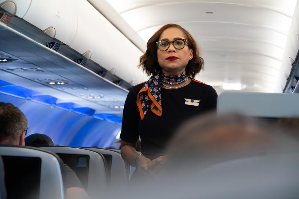 A flight attendant wearing glasses and a patterned neck scarf stands in the aisle of an airplane with passengers seated