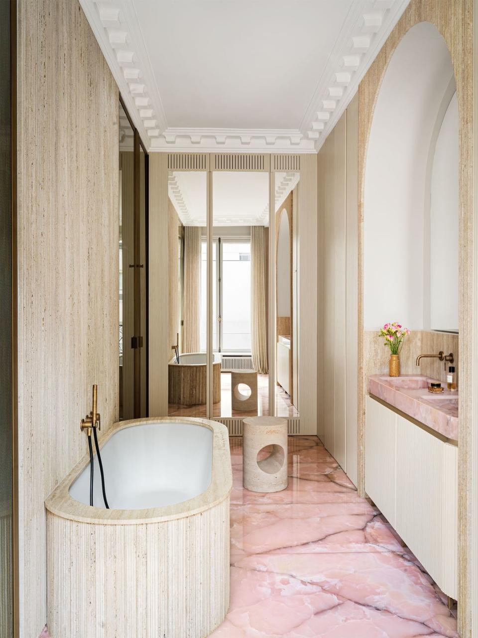 bathroom with pink marble floor and sink top, bathtub with grooved slats on exterior, cylindrical side table or stool, mirrored wall, sinks inset into arched wall, grooved lower cabinets