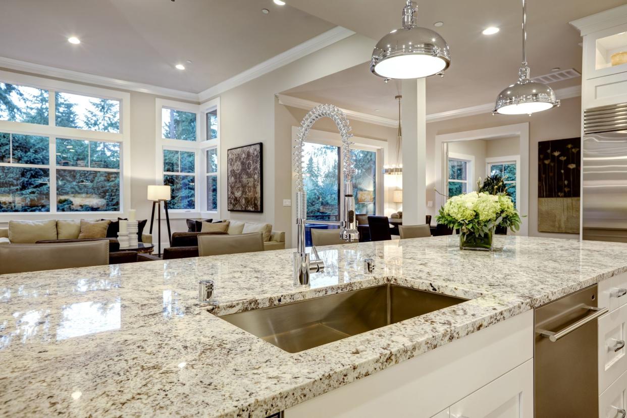 White modern kitchen featuring island with granite countertop, island has sink, living room and two other rooms in the background