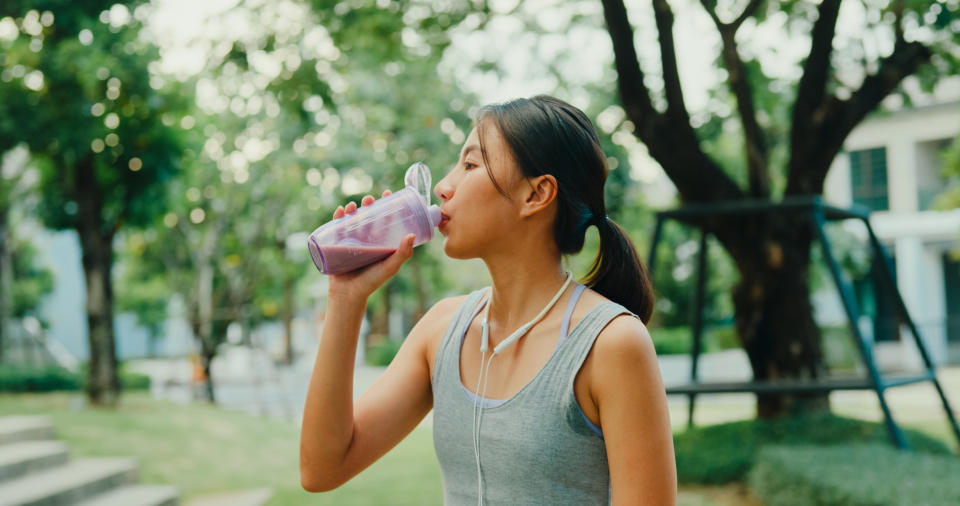 A woman drinks from a water bottle while standing outdoors in a park, wearing workout attire and earphones