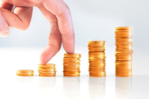 A person's fingers climbing a series of gold coins stacked in columns that increase in size.