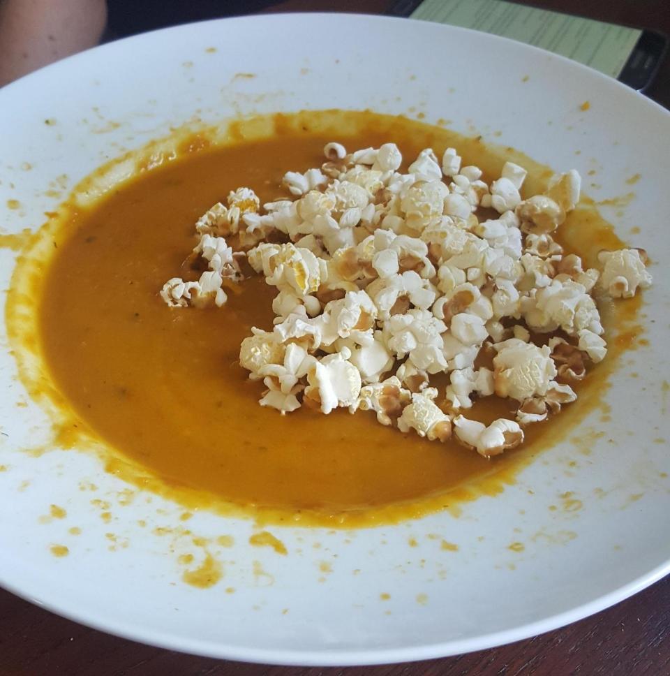 A bowl of creamy soup topped with popcorn sits on a white plate