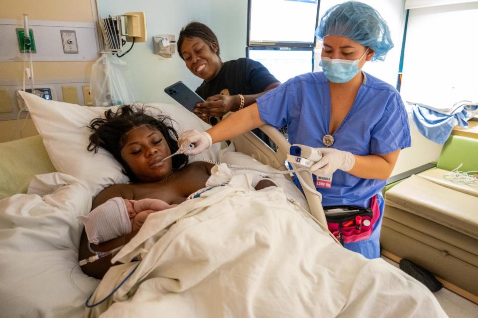 A woman takes a photo of a woman in bed holding a baby as a nurse attends.