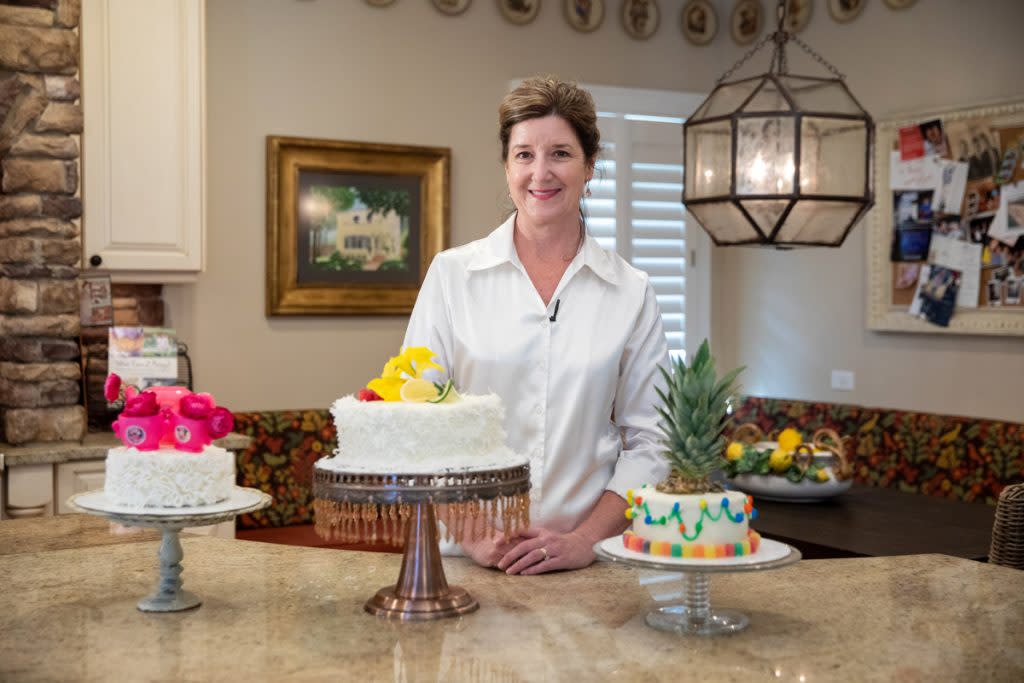 A woman smiles in this portrait by three cakes she made. Two of them have fondant. One of those two is decorated with fresh fruit and the other is decorated with a plastic toy Minnie Mouse tea set. The third cake is decorated with candy and the top of a pineapple. 