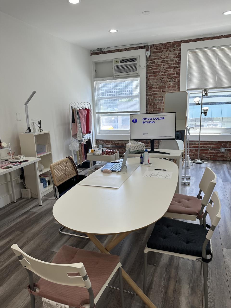 A modern, well-lit office with a white table, chairs, computer monitor displaying "ONYO COLOR STUDIO," various office supplies, and a clothes rack in the background