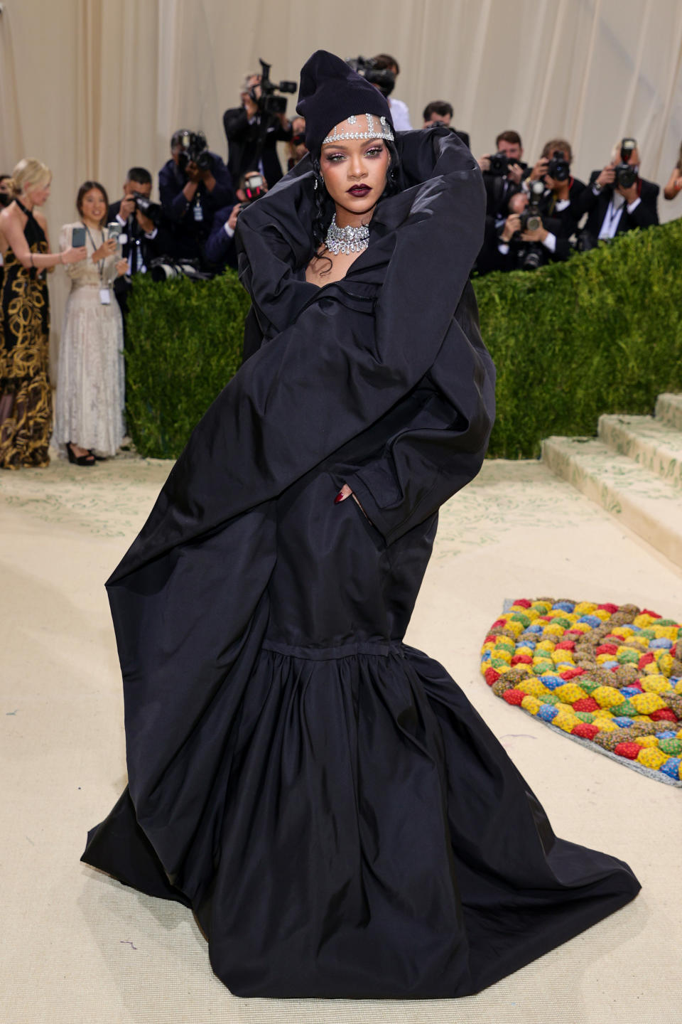 Rihanna poses on the red carpet in a voluminous dark-colored gown, a beanie, and a large diamond necklace