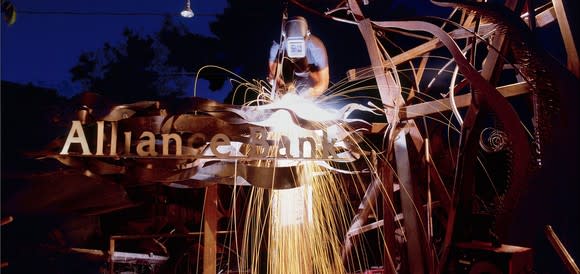 Welder working on metal structure in front of Alliance Bank logo.