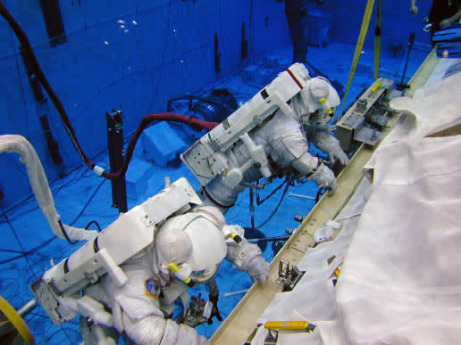 Two astronauts in spacesuits are seen underwater, holding onto a railing.