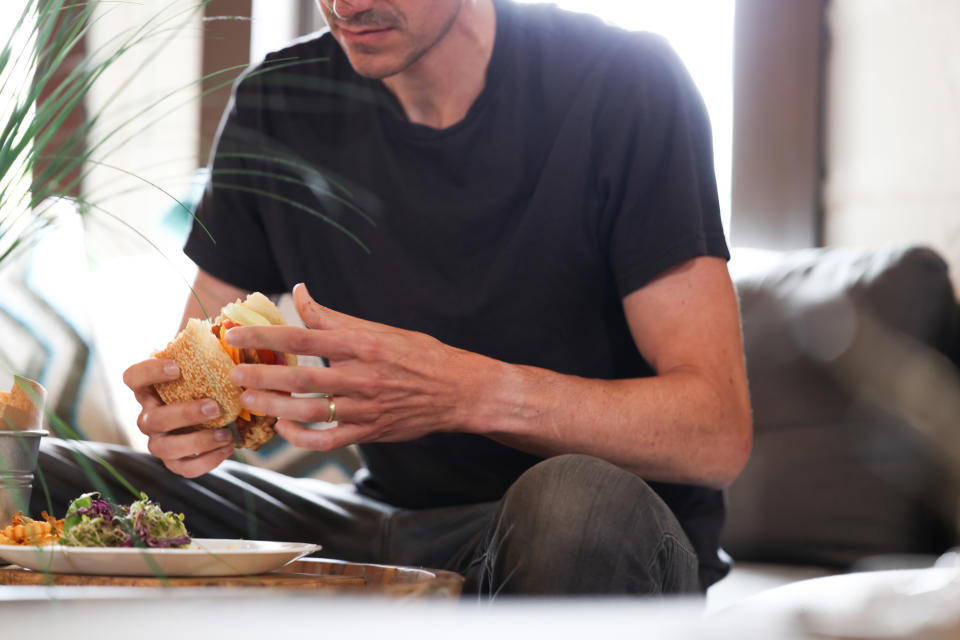 A man sits on a couch, holding a sandwich close to his mouth, appearing ready to take a bite. A plate of food is on the table in front of him