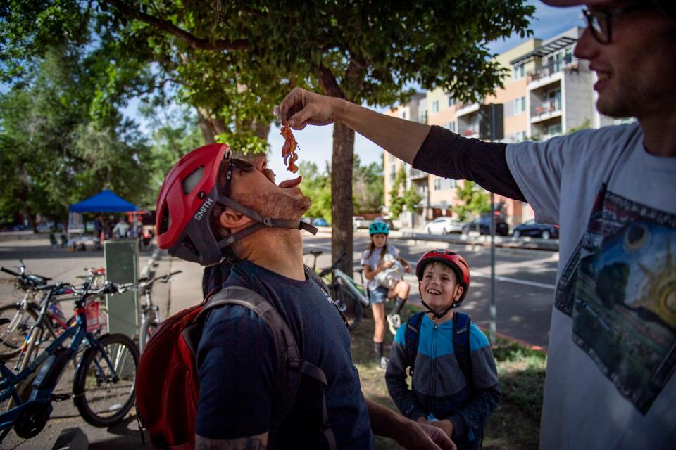 Wes Wickland is fed a piece of bacon by Milan Krucky at the Recycled Cycles booth on South Mason Street during Bike to Work (or Wherever) Day in Fort Collins on June 22, 2022.