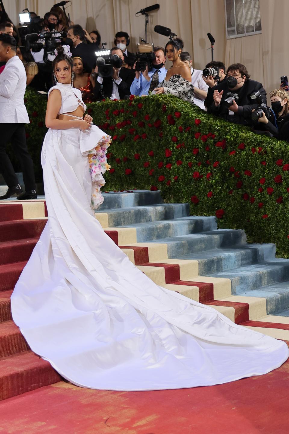 Camila on the red carpet steps with a long white skirt train flowing down them. The front of the skirt has a large ruffle with pastel-colored flowers within the ruffle. She has a crop top that reveals her bare torso and sternum below her chest with white straps wrapped around her stomach. She has her hair up in a bun.