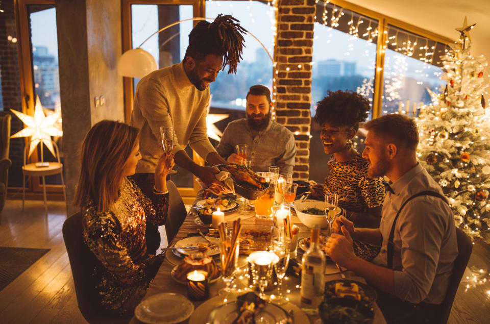 People sitting at a festive dinner table smiling, chatting, and raising glasses in a warm, decorated room with lights, a star lamp, and a Christmas tree in the background