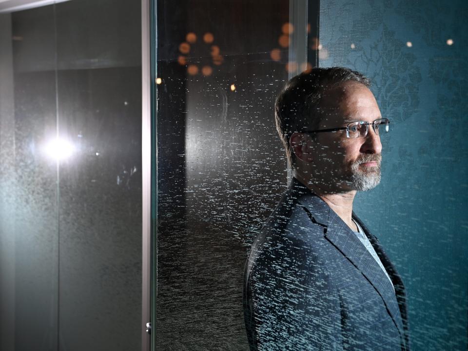 A man with grey hair, beard, and glasses stands behind a glass wall staring off into the distance.