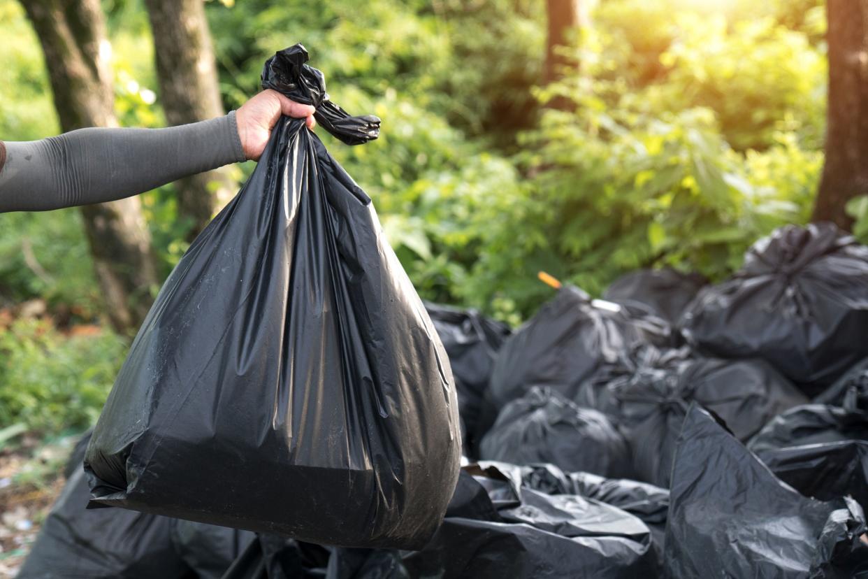 hand holding large black trash bag outside with trash pile of other trash bags and woods in the background