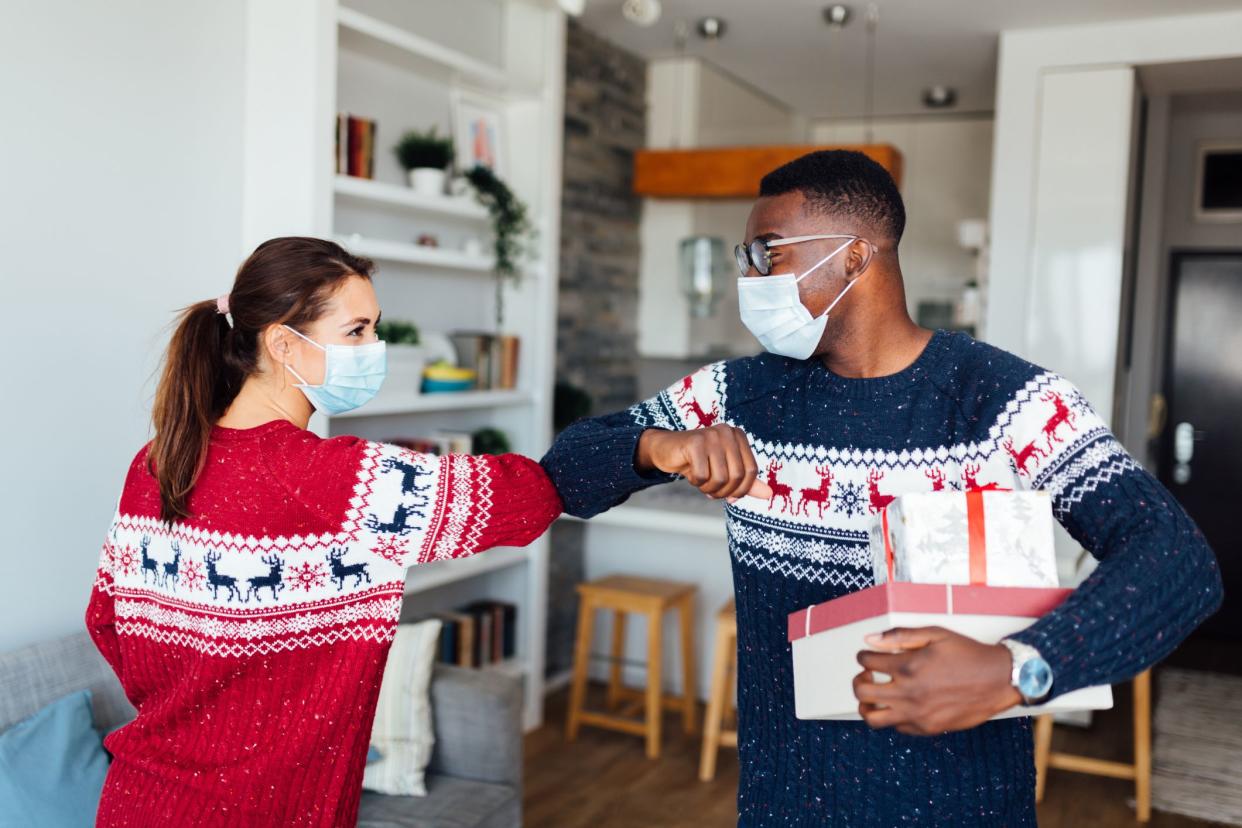 Young Caucasian woman having her friend of African American ethnicity in her home for winter holidays, bringing gifts, wearing protective face mask, keeping social distancing and elbow bumping to greet each other