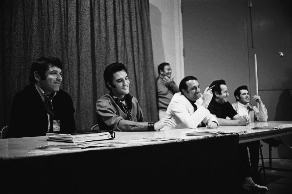 a black and white photo of five men, including elvis presley, sitting at a long table during a press conference and smiling