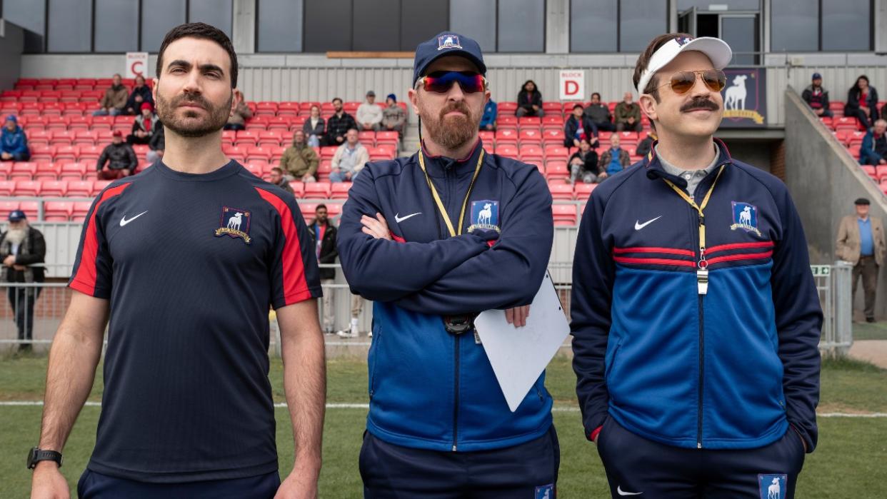  From left to right: Brett Goldstein, Brendan Hunt and Jason Sudeikis standing on the pitch in Ted Lasso. 