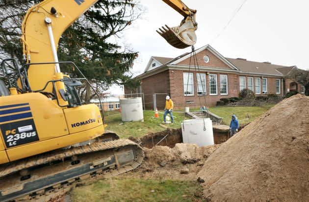 Workers use heavy machinery to lower a section of concrete pipe into the ground by a school district building in Wyandanch, a hamlet on Long Island, on Dec. 15, 2021. The work is part of a project to connect the building to the sewer system.