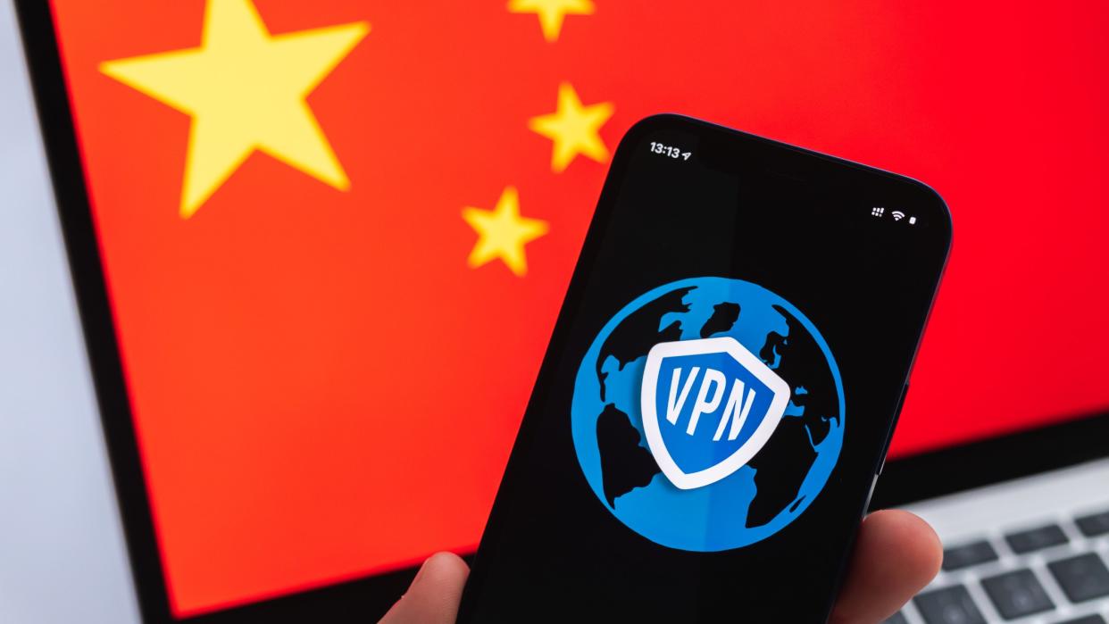  A hand with mobile phone and VPN app in the foreground, and a Chinese flag on a laptop screen in the background 