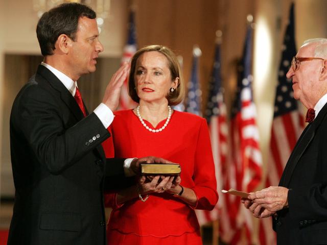 A photo of John Roberts raising his right hand while his left sits on a Bible during his 2005 swearing in as Chief Justice of the Supreme Court. His wife, Jane Roberts, stands next to him.
