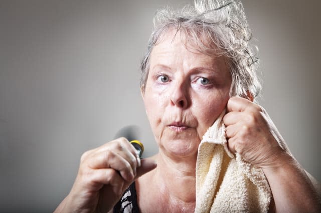 woman in her fifties sweatening,  having hot flashes and trying to cool with small fan, drying face with towel