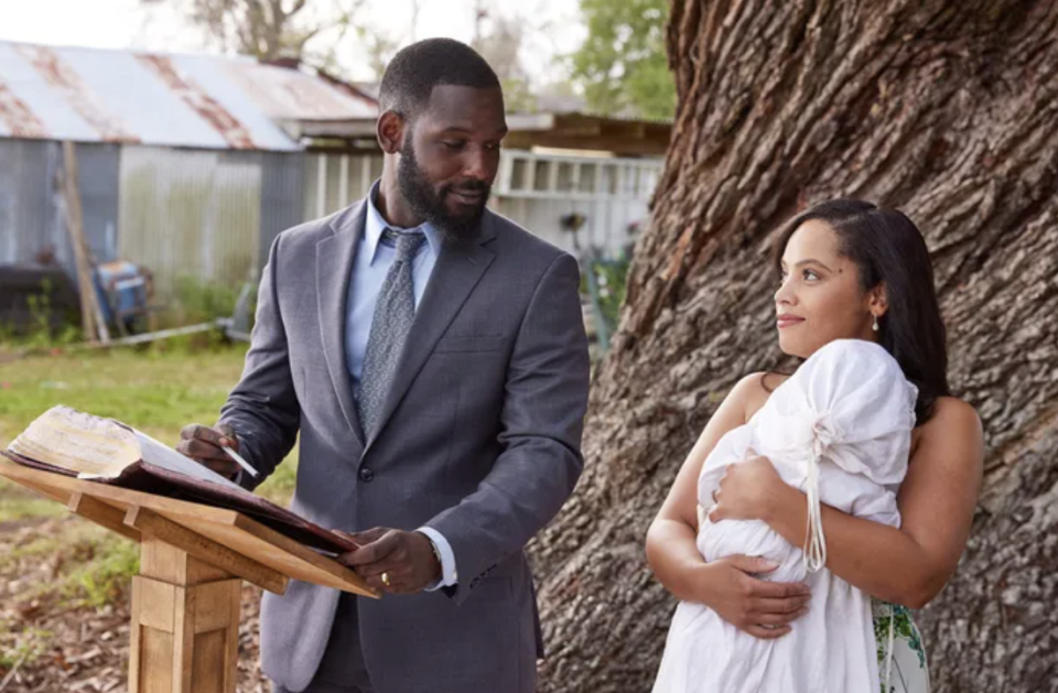 Man in a suit stands by a tree with a book on a stand; woman in a draped gown holds an infant, both looking at each other