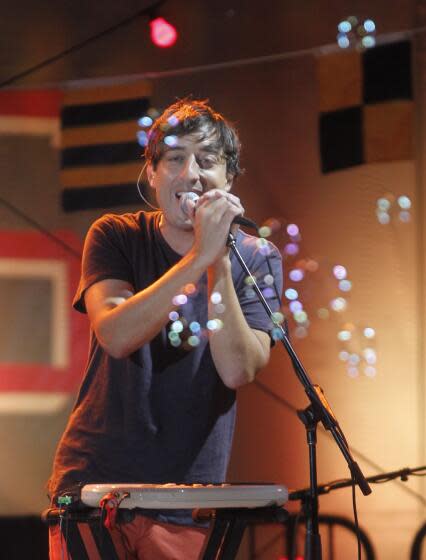 Ed Droste singing into a microphone behind a window in a recording studio