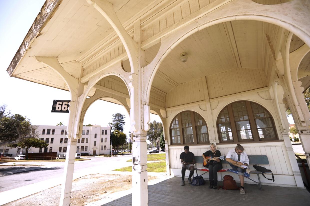 Three people sit on a bench outside a white building.