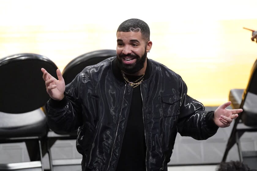 A man with a beard wearing a black shirt and black bomber jacket holds his hands up