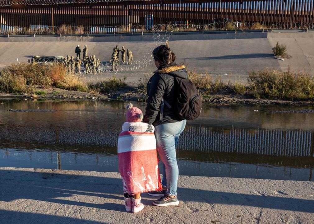 View of woman and child from behind on Ciudad Juarez side of border with border agents visible across the river. 