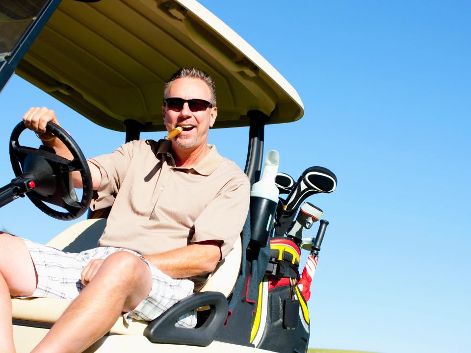 Older man smoking a cigar while driving a golf cart on a sunny day.